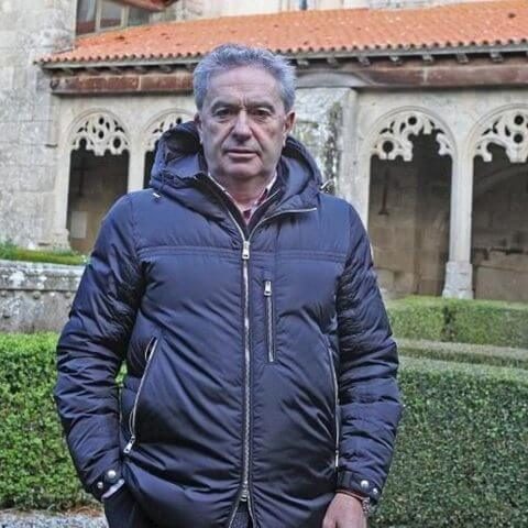 Retrato do alcalde de Xunqueira de Ambía, vestido cun chaquetón escuro de abrigo, de pé no xardín do claustro da colexiata cos arcos de pedr
