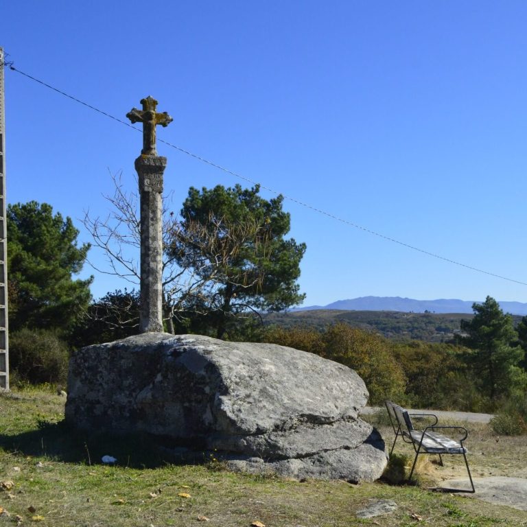 Cruceiro de pedra tradicional asentado sobre unha gran rocha natural, situado nunha contorna verde con árbores e un banco de madeira ao lado