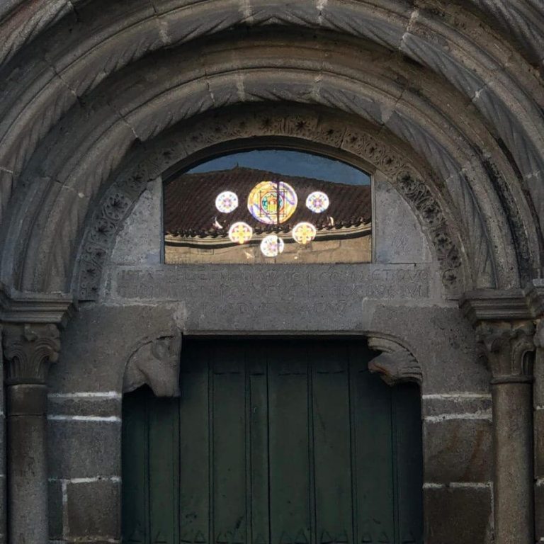 Porta principal pechada, enmarcada por un groso arco de pedra, que ten xusto encima unha pequena ventá circular decorada con vidreiras.
