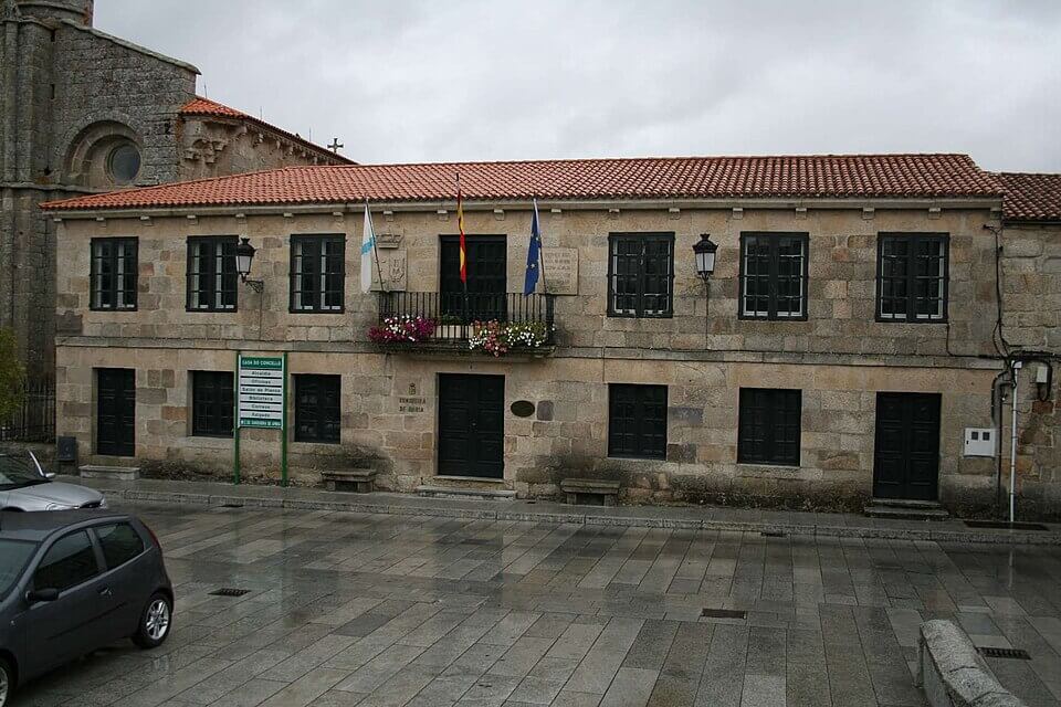 Concello Fachada principal da casa consistorial de pedra con dous pisos, portas e ventás simétricas de madeira escura e tres bandeiras oficiais no ba