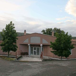Fachada simétrica dun edificio moderno dunha soa planta con paredes de cor salmón, zona central acristalada e dúas árbores a cada lado.