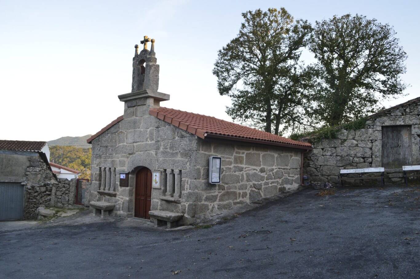 Capela San Xillao Pequena capela tradicional de cantería cunha espadana no tellado, porta de madeira e unha ventá estreita lateral, rodeada de árbores.