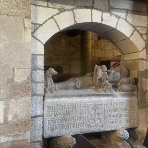 Sepulcro antigo de pedra incrustado nun nicho da parede baixo un arco, coa estatua dunha persoa xacente sobre unha caixa con inscricións.