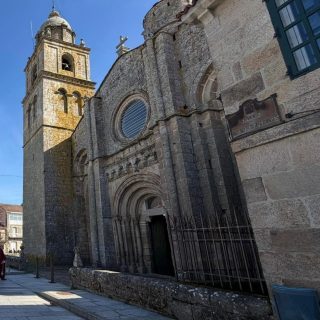 Vista exterior da fachada románica de pedra da colexiata, coa porta de arcos de medio punto, contrafortes e a torre do campanario ao fondo.