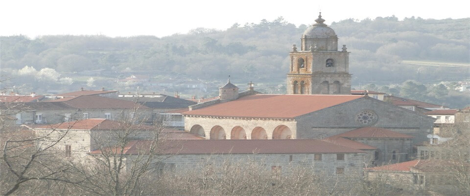 Panorámica da vila Paisaxe xeral da vila nun día de brétema, onde destacan construcións de pedra con arcadas e a alta torre do campanario no horizonte.