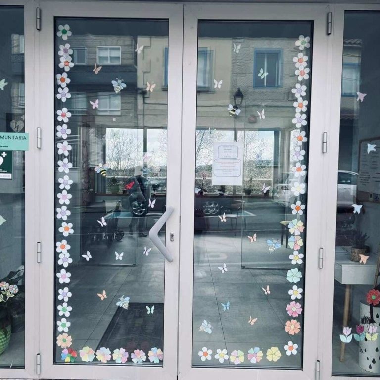 Porta principal de dobre folla de cristal, decorada con pegatinas de pequenas flores de cores, mostrando o interior iluminado do centro.