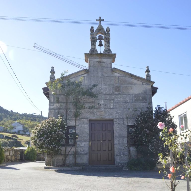 Capela rústica cunha cruz de pedra no alto e outra cruz sobre a porta principal, con canteiros de flores ás beiras da entrada.
