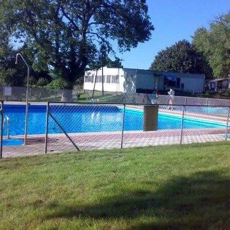 Vista das instalacións das piscinas municipais ao aire libre, mostrando o vaso de auga azul rodeado por un valado metálico e zona de césped.